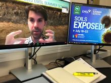 A pen, a notebook, and two computer monitors are showing. One monitor displays a male presenter gesturing with his hands as he speaks. The other monitor display text: "Soils Exposed! July 18-22."