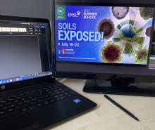 A pen, laptop computer, and monitor with screen text [Soils Exposed! July 18-22] 