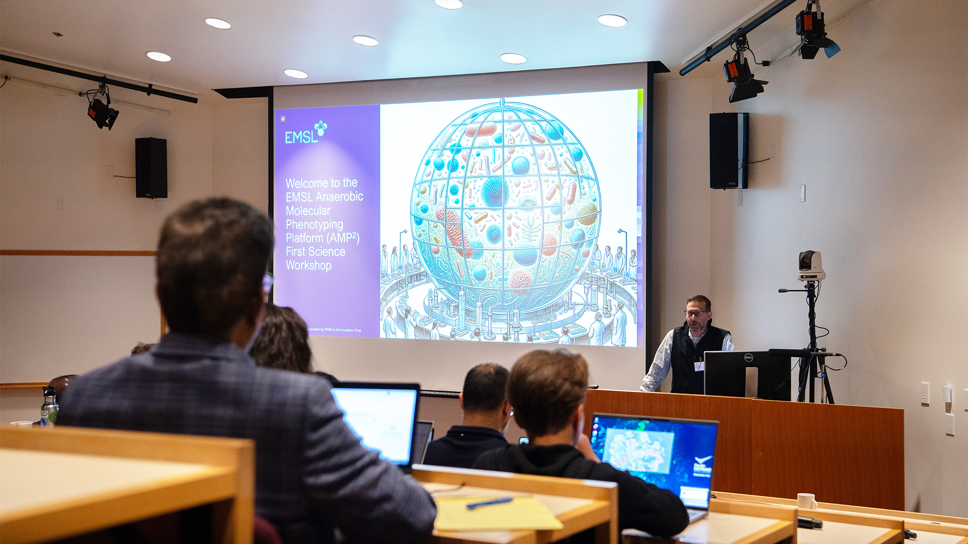 Photo of researcher Scott Baker presenting, standing in front of screen with the following words projected—Welcome to the EMSL Anaerobic Molecular Phenotyping Platform (AMP2) First Science Workshop.