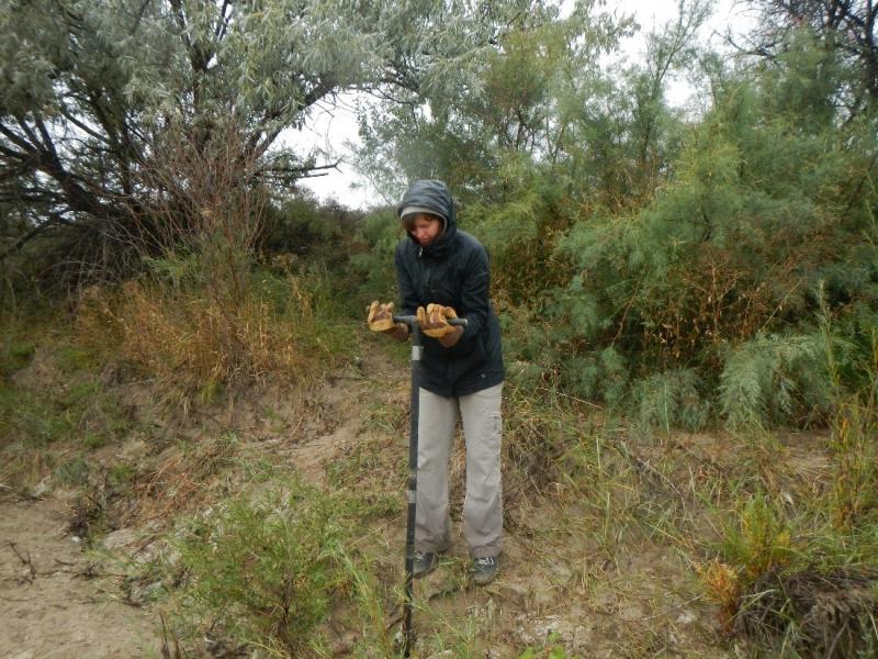 person in hooded jacket holds T-shaped device in dirt.