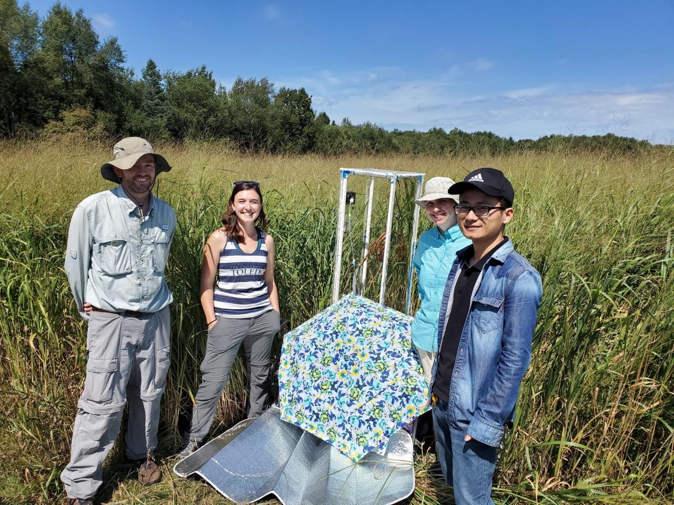 Four people standing in a field with tall grasses. Umbrella and sun shade are in the foreground.