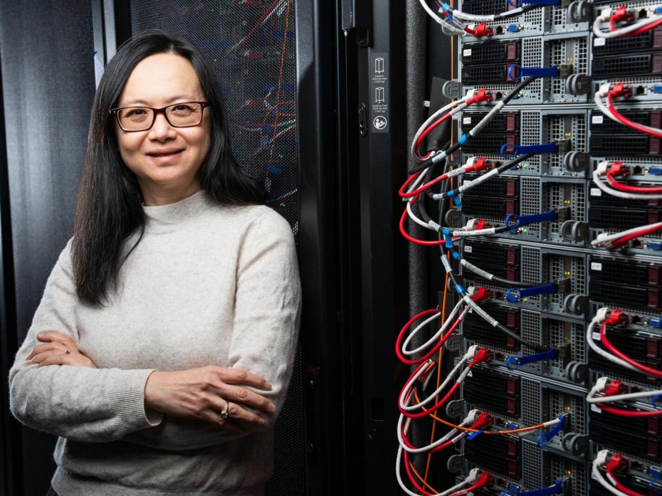Photo of researcher Margaret Cheung smiling at the camera, standing next to a wall of computing equipment.